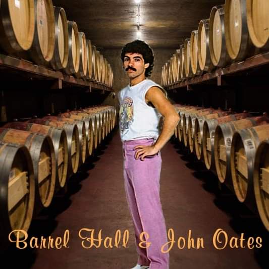 A man with a mustache and curly hair stands confidently in a wine cellar aisle flanked by rows of wooden barrels. He wears a…