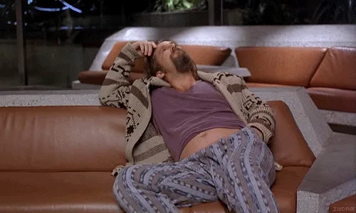 The image shows a man in pajamas lying on a brown couch with his head tilted back and hands on his head, appearing exhausted…