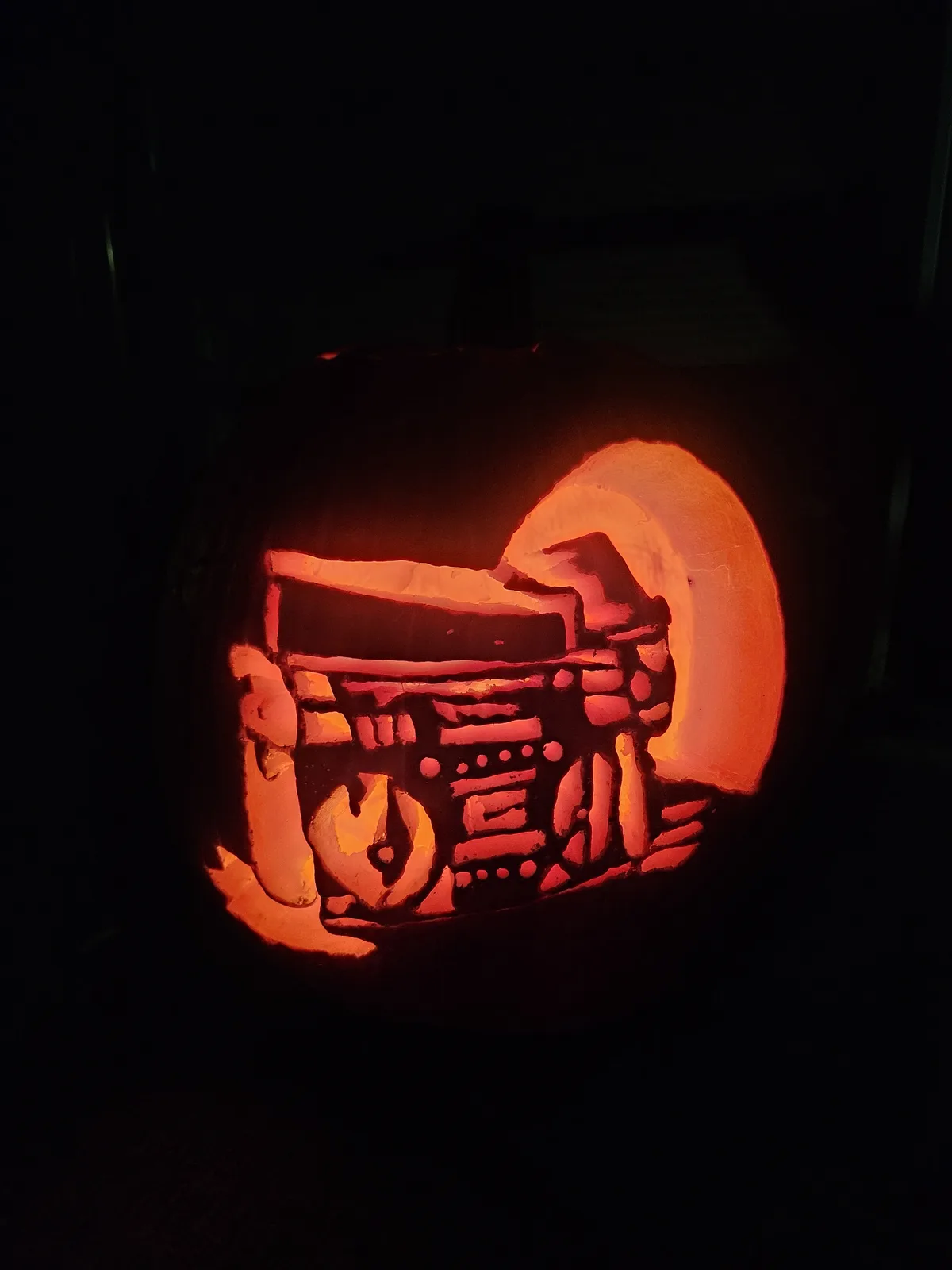 A carved pumpkin illuminated from within, featuring a detailed design of a boombox with a hand holding a cassette tape, set…