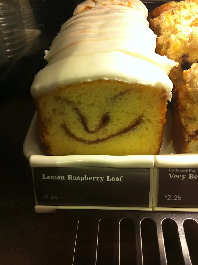 A close-up of a lemon raspberry loaf cake in a bakery display case, topped with white icing and featuring a smiling face…