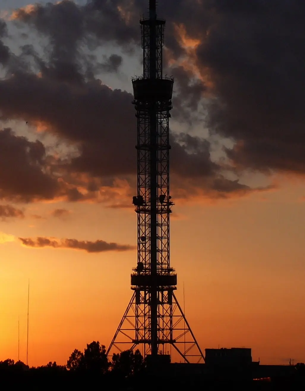 A tall, silhouetted communication tower stands against a dramatic sunset sky with orange and dark clouds. The image has text…