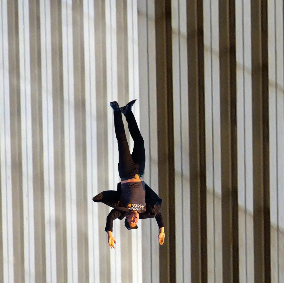 A man is suspended upside down against a wall with vertical stripes, wearing a black outfit and a cap, with his arms…