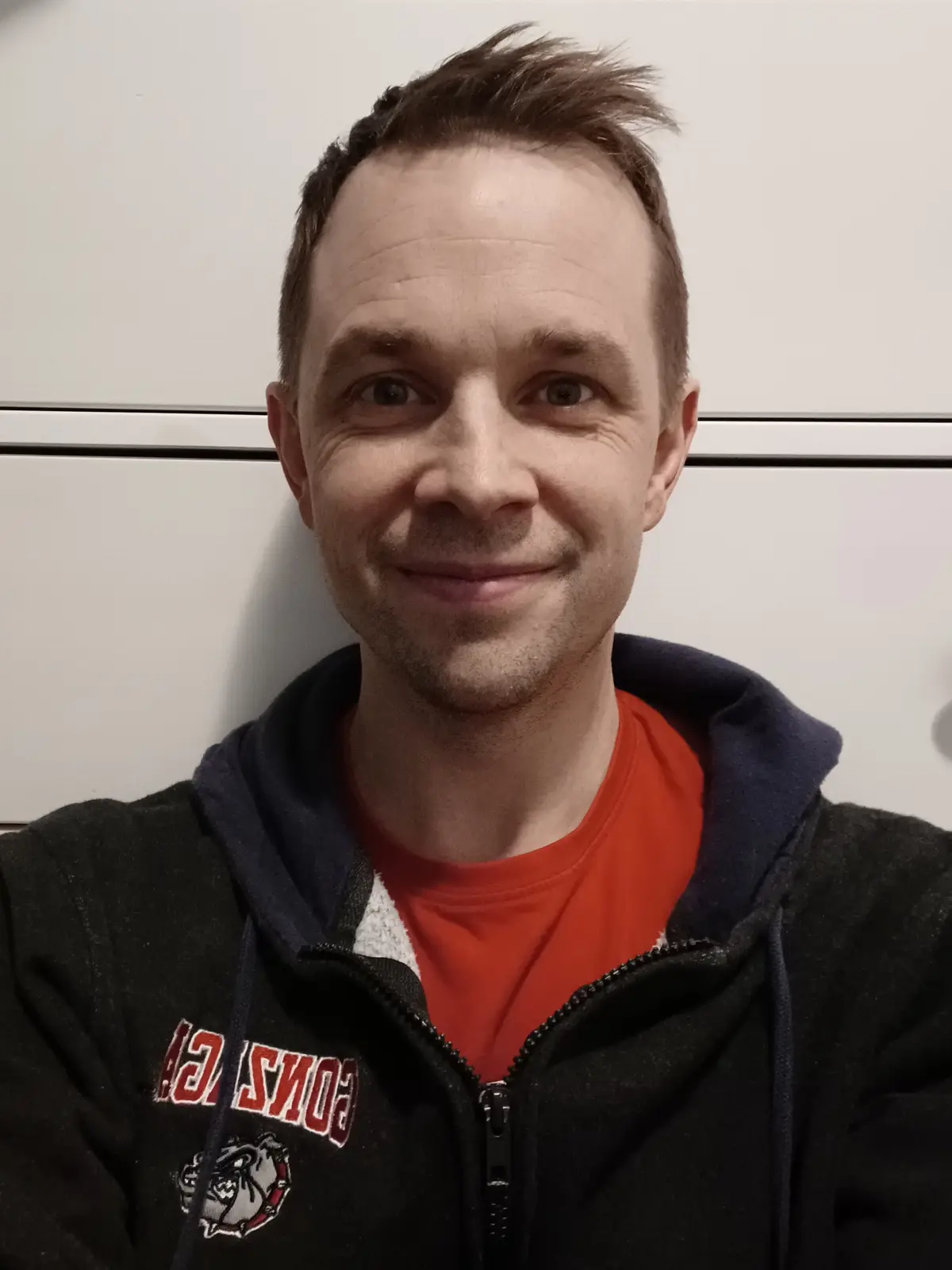 A man with short brown hair and a slight smile wears a black zip-up hoodie over an orange t-shirt, standing in front of…