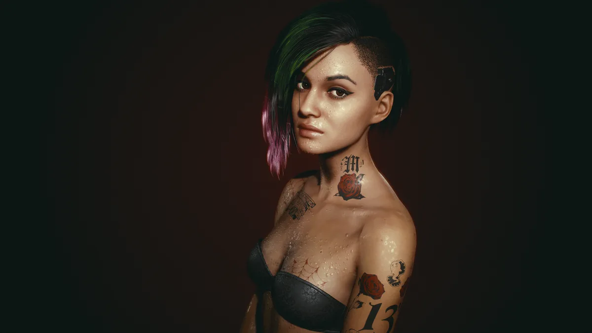 A woman with short, dark hair streaked with green and purple, wearing a black bra, has multiple tattoos including a rose and…