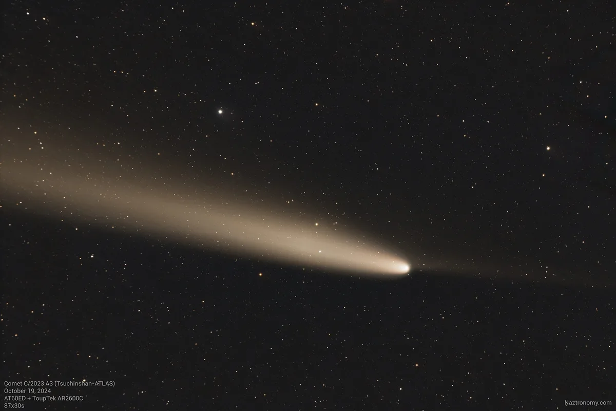 A celestial photograph shows a bright comet with a long, diffuse, beige-colored tail stretching diagonally across a…