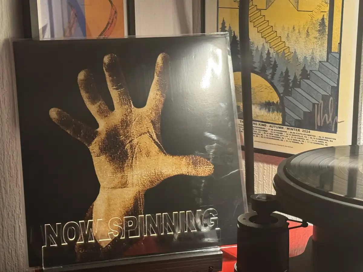 A vinyl record sleeve featuring a large, textured golden handprint against a black background, with the text 'NOW SPINNING'…
