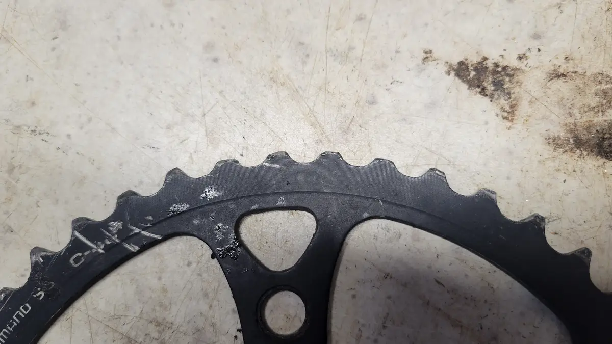 A close-up photo of a black, worn bicycle chainring with visible scratches and white residue on its teeth, resting on a…