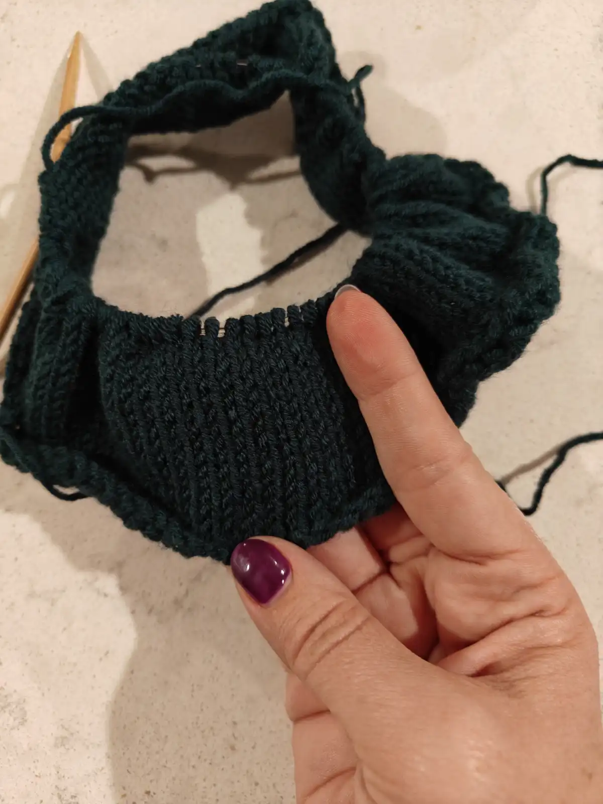 A hand with purple nail polish holds up a dark green, circular knitted item that resembles a donut with a hole in the…