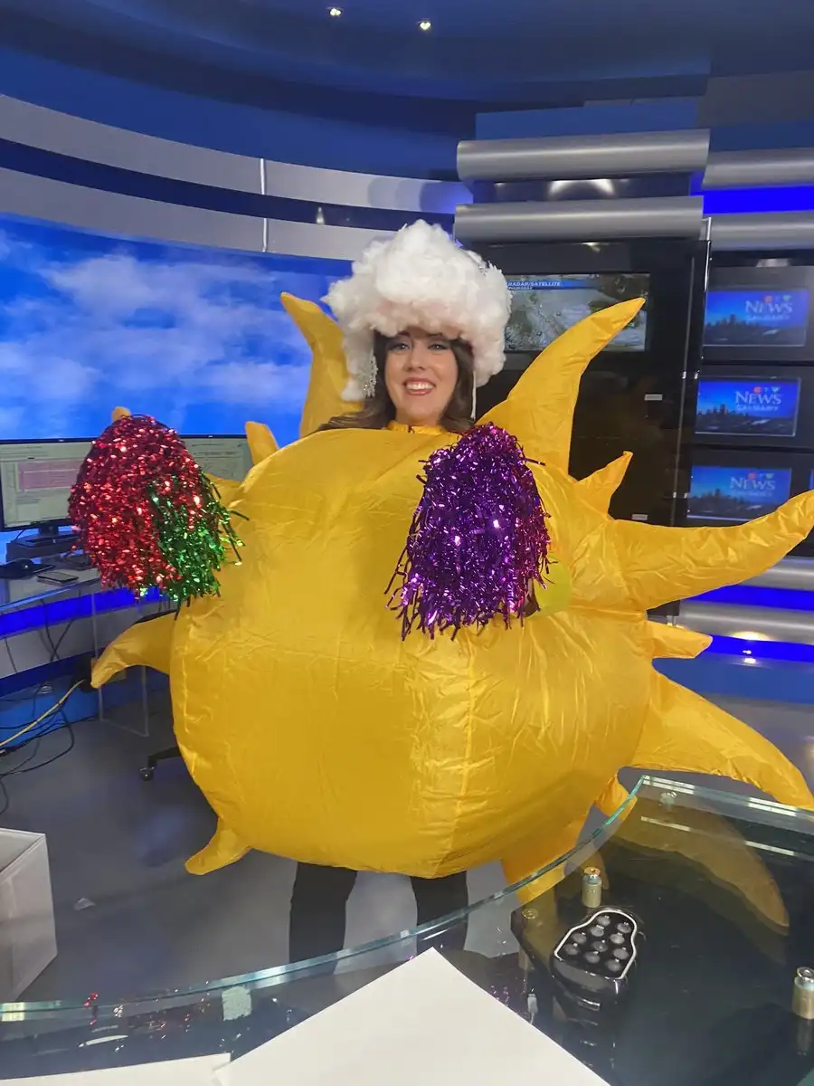 A woman in a news studio wearing a large, inflatable yellow sun costume with white fluffy hat, holding red and purple…