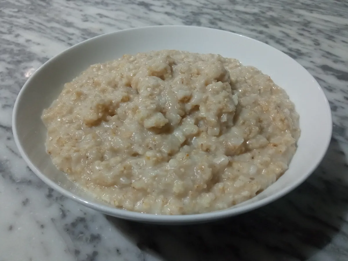 A close-up photo shows a white ceramic bowl filled with creamy, lumpy oatmeal resting on a gray and white marble countertop…