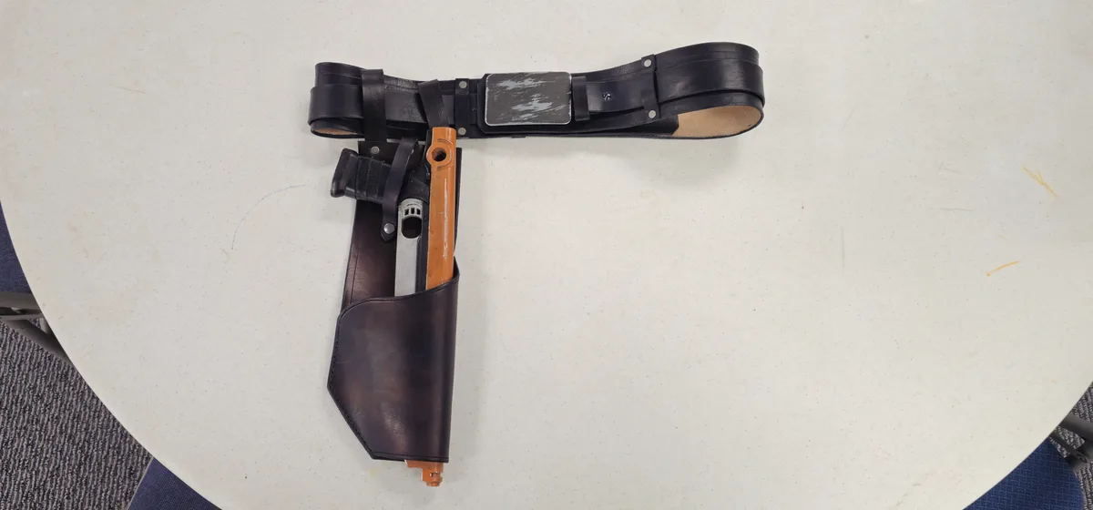 A black leather belt with a silver buckle and a matching holster containing a toy blaster with an orange grip lies on a…
