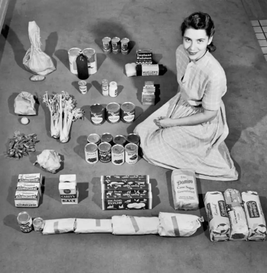 A black-and-white photo shows a smiling woman in a 1950s-style dress kneeling on a floor surrounded by a large assortment of…