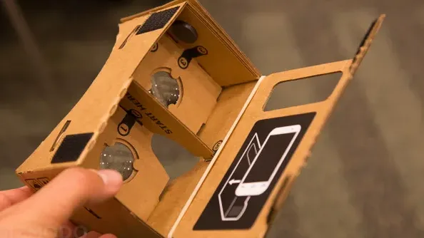 A hand holds a cardboard Google Cardboard VR headset, showing its interior lenses and a printed diagram on the side…
