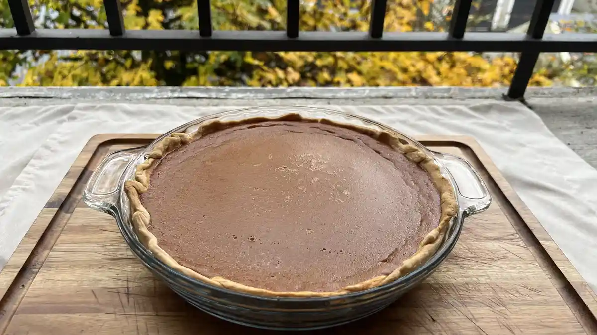A freshly baked chocolate pie sits in a clear glass dish on a wooden cutting board, placed on a white cloth over a balcony…