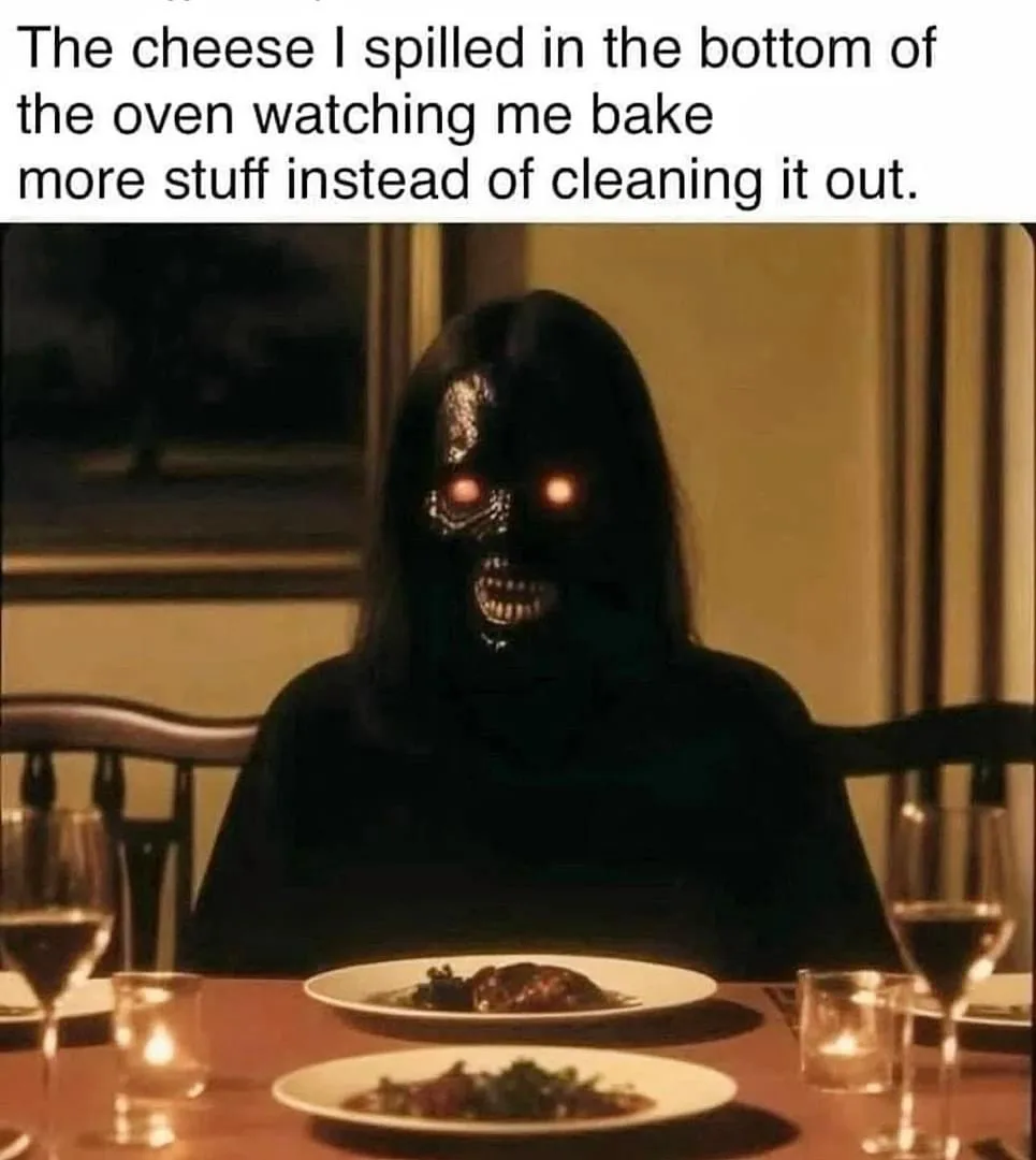A person wearing a black hooded costume with a skull mask featuring glowing red eyes sits at a dining table set with plates…