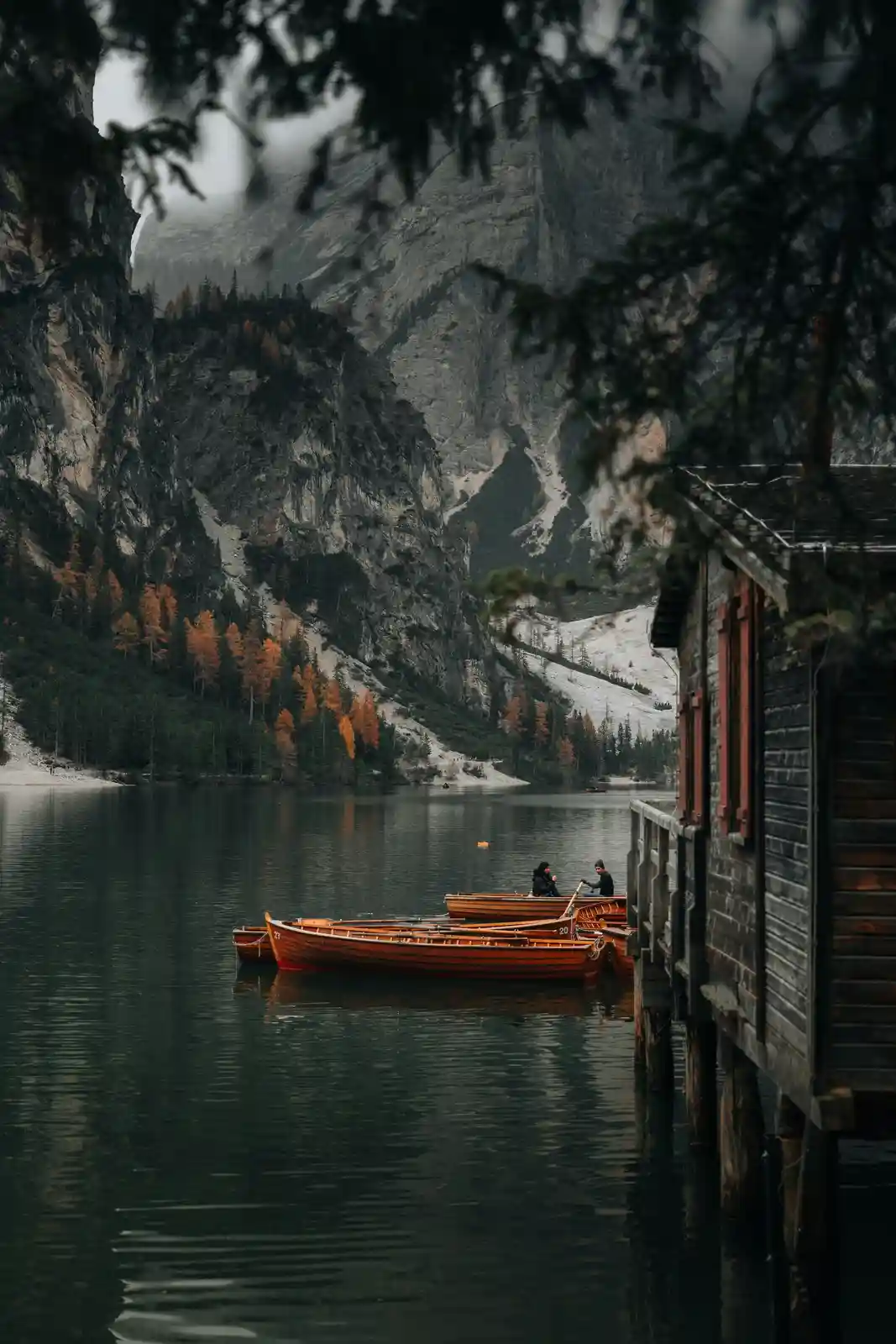 A serene, moody landscape photo shows a calm lake surrounded by towering, misty mountains with autumn-colored trees. Several…