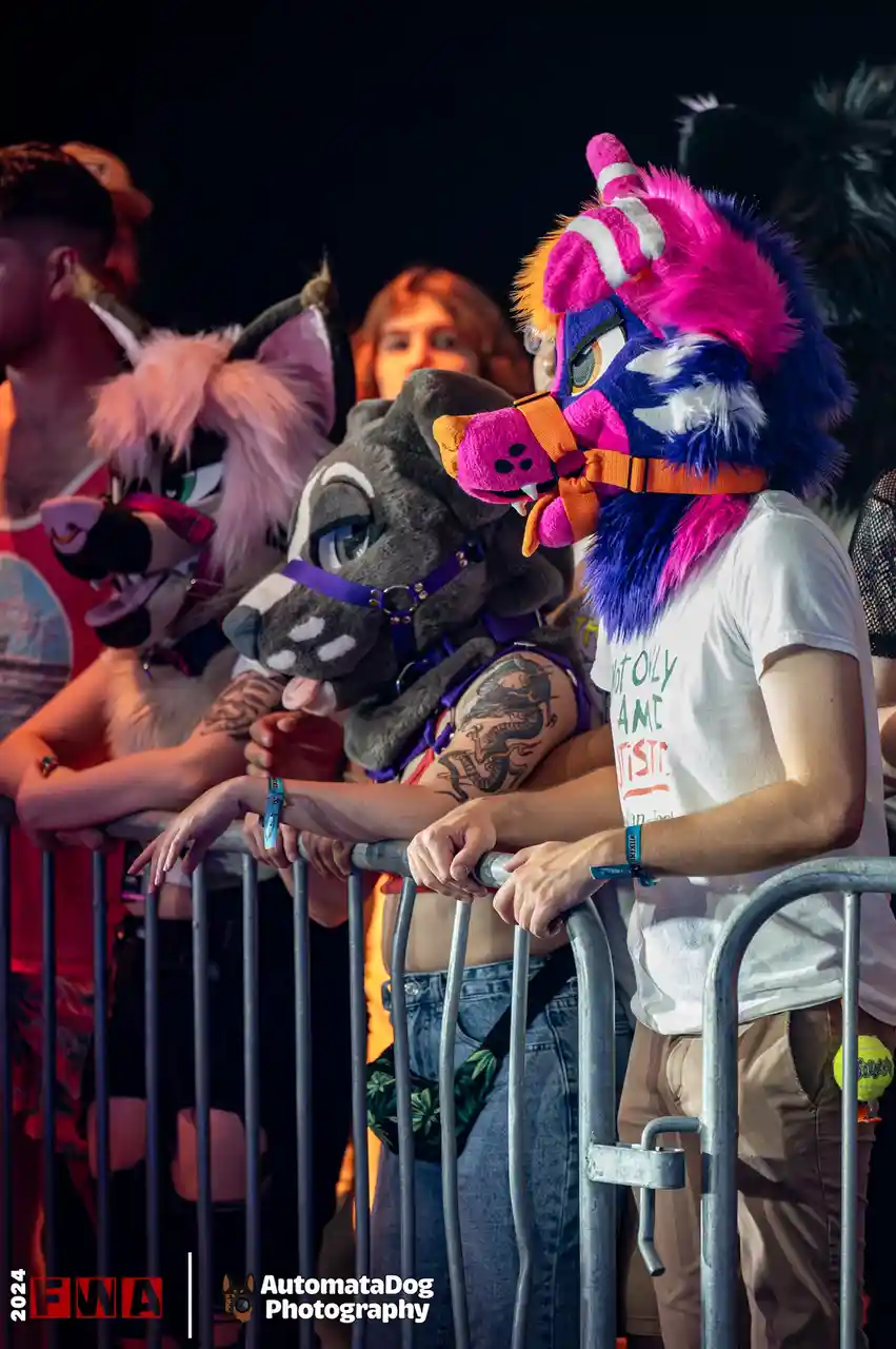 Several people in colorful, elaborate fursuit costumes lean on a metal barrier at a nighttime event. The person on the…