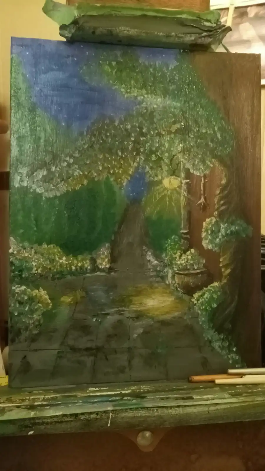 A painting on an easel depicts a garden scene with trees, bushes, and a stone path under a blue sky, with visible…