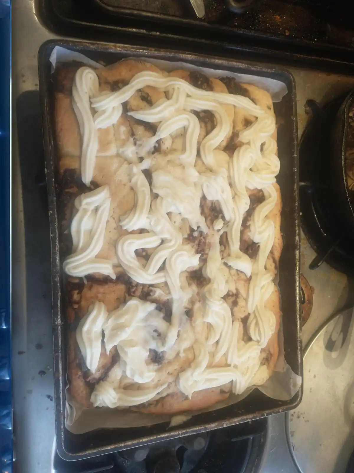 A rectangular baking pan holds a batch of homemade cinnamon rolls, golden-brown and topped with thick, white icing piped in…