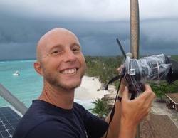 A bald man in a dark t-shirt smiles broadly while holding a DSLR camera with a plastic bag covering the lens, standing on a…