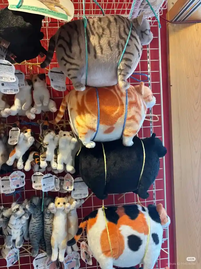 A photo shows several large, plush cat keychains hanging from a wire rack in a store, arranged in a stack with their…
