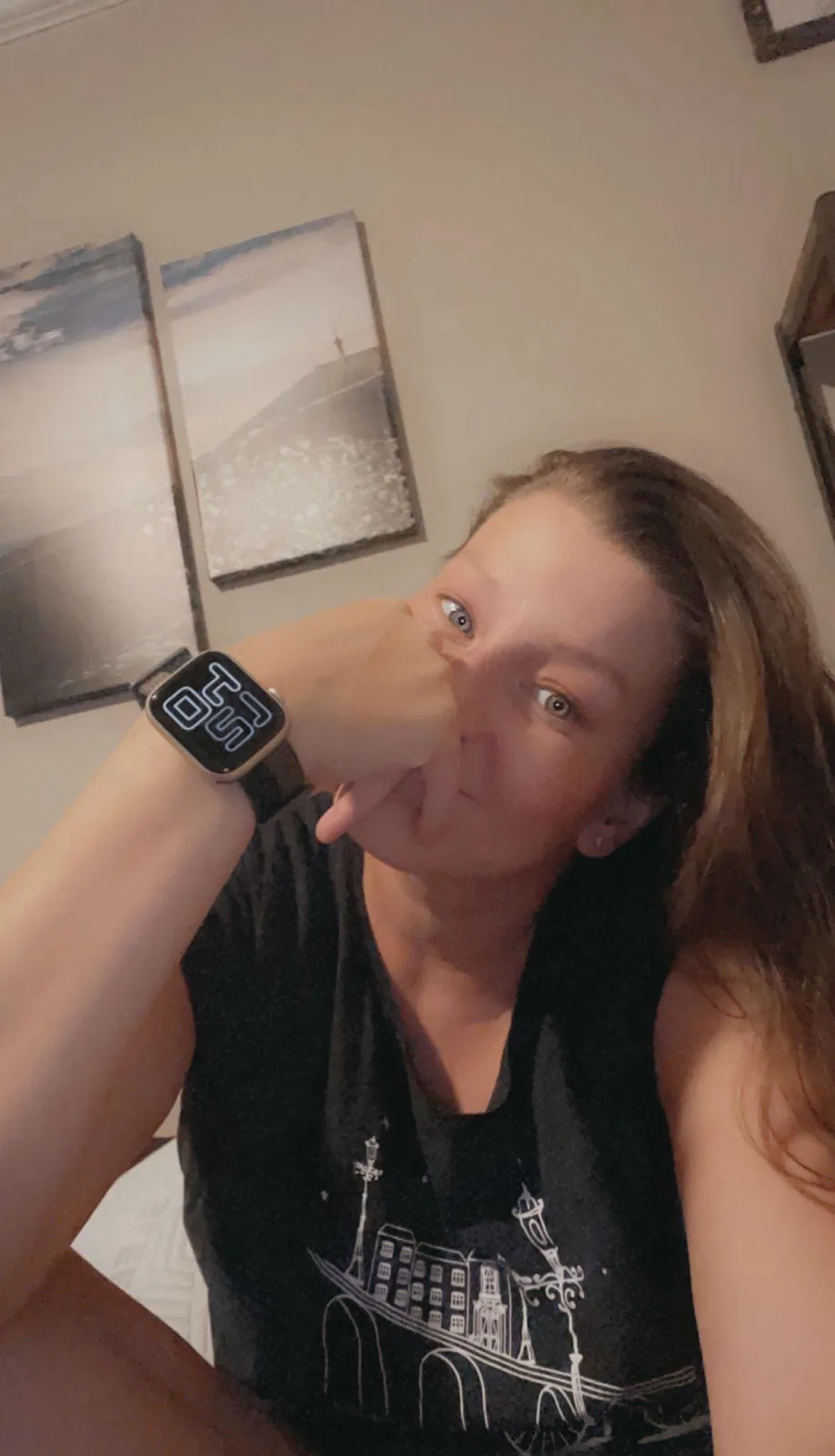 A person with long brown hair wearing a black tank top takes a selfie, holding their hand over their mouth and nose while…