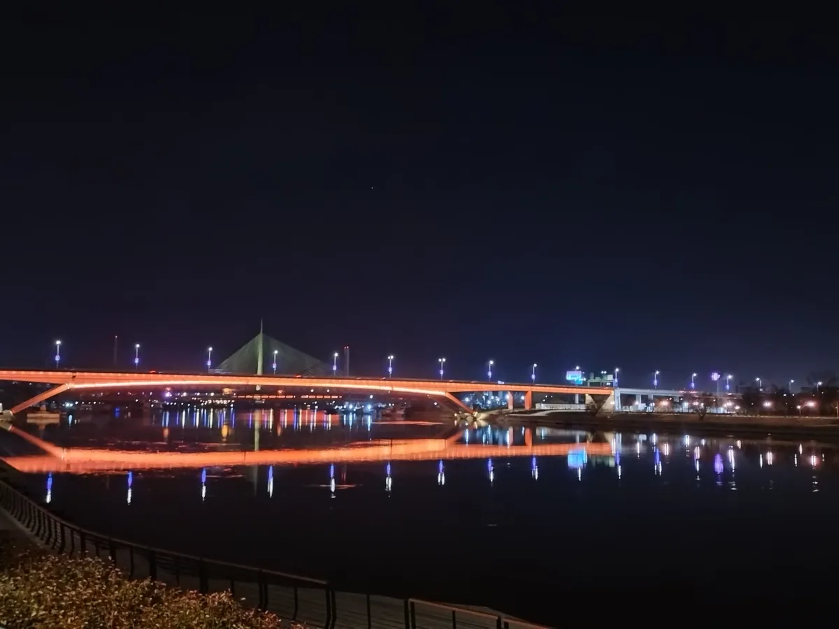 A nighttime photo of a brightly lit orange bridge spanning a calm river, with its lights reflecting in the water. A modern…