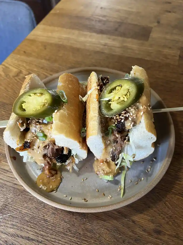 A close-up photo shows a sandwich cut in half, served on a ceramic plate on a wooden table. Each half is topped with a slice…