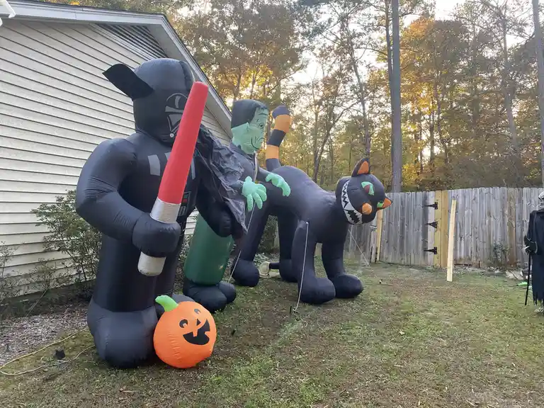 An outdoor Halloween scene shows three large inflatable decorations: a Darth Vader figure holding a red lightsaber, a green…