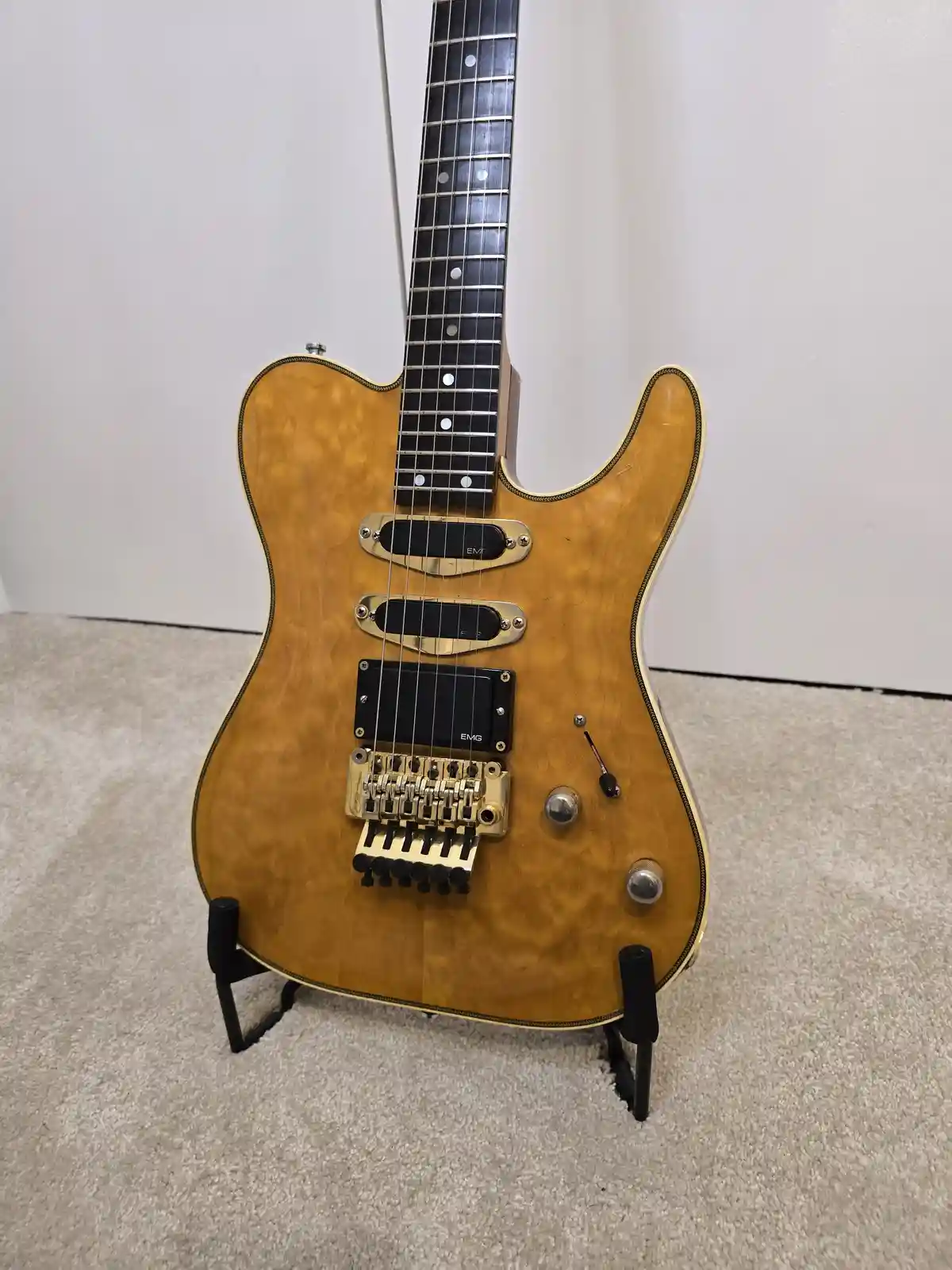 a light-wood electric guitar with a visible EMG pickup and gold hardware stands upright on a black stand against a plain…