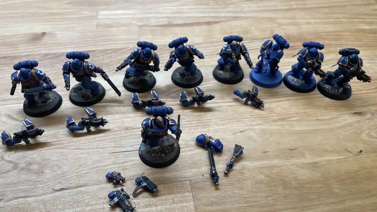 A row of blue-painted Space Marine miniatures stands on a wooden floor, surrounded by their disassembled weapons and limbs…