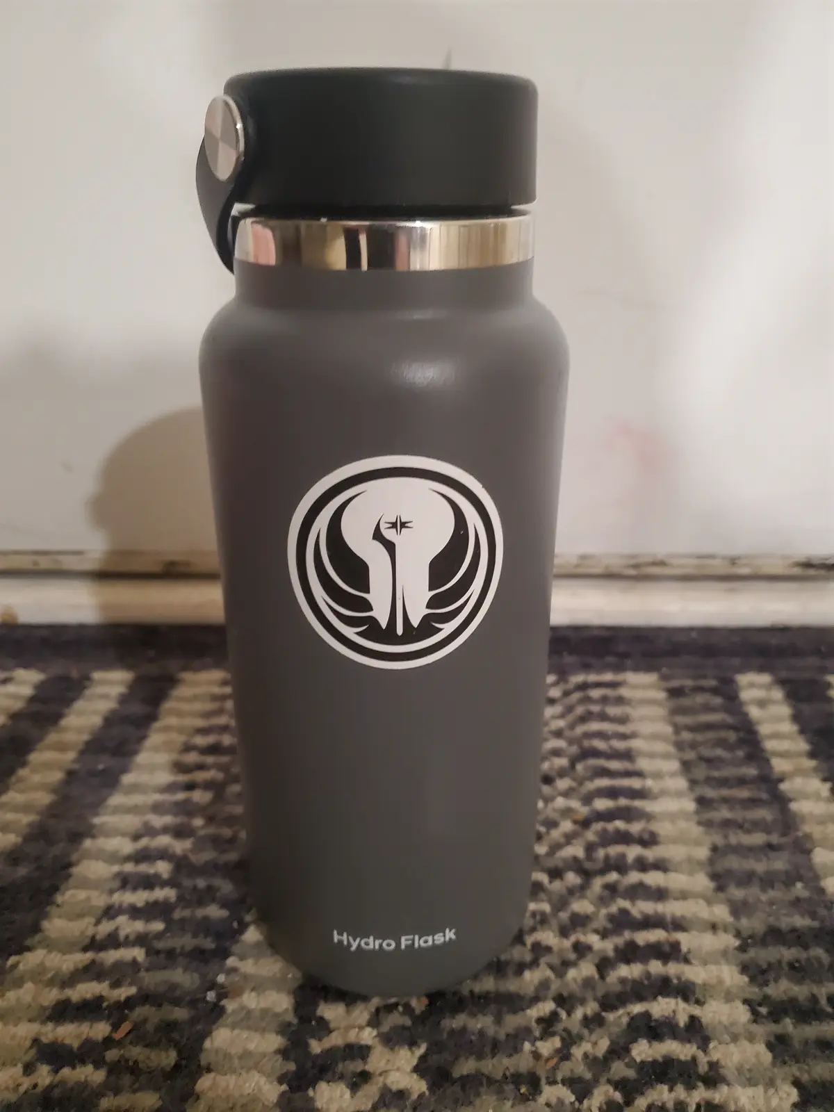 A matte gray Hydro Flask water bottle with a black lid and silver band sits on a patterned rug. A white circular sticker on…
