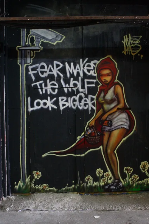 A street mural depicts a woman in a red hooded cloak holding a basket, standing beside a lamppost with a security camera…