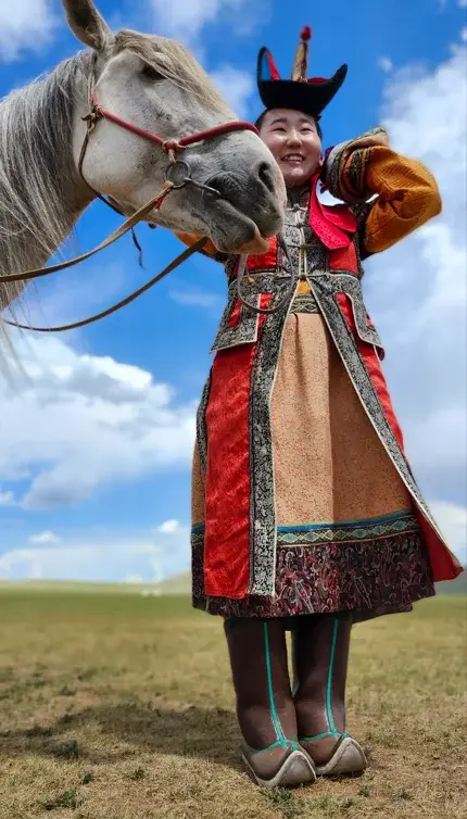 A person in traditional ornate red and gold clothing stands in a grassy field under a blue sky, smiling while a white horse…