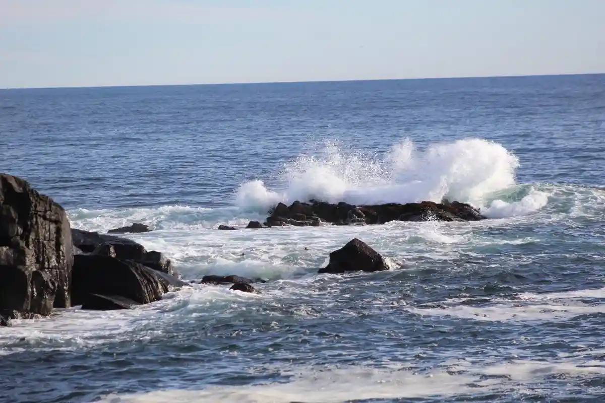 A photograph of ocean waves crashing violently against dark, jagged rocks, creating white foam and spray. The overlaid text…