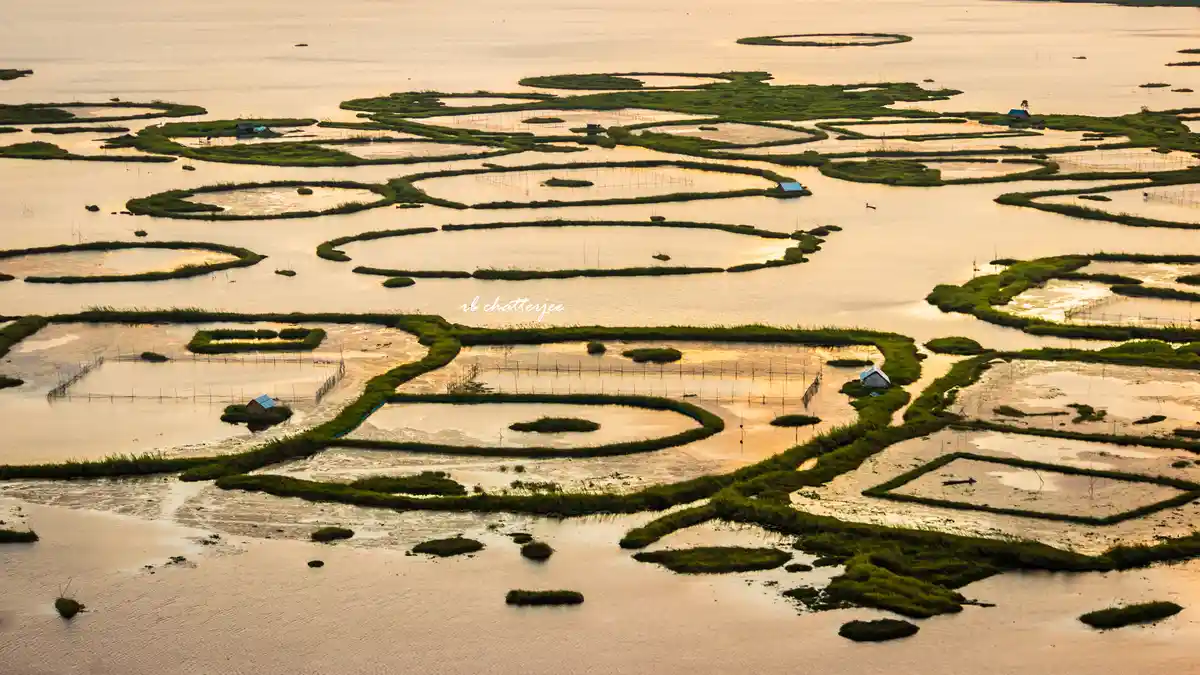 An aerial view of a flooded landscape with green grassy islands and water channels, resembling a complex maze or circuit…