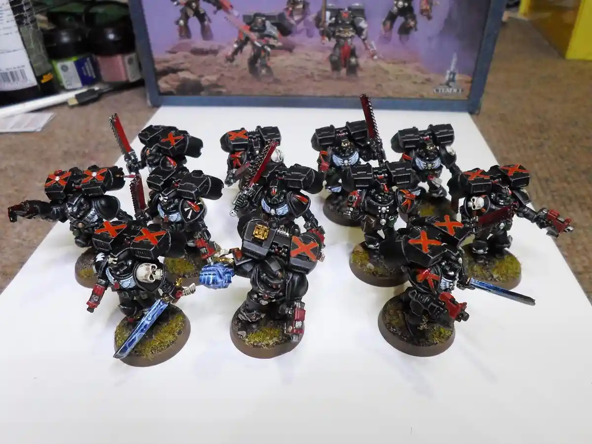 A photo of several painted miniature warhammer 40k space marines, mostly black with red crosses on their armor, standing on…