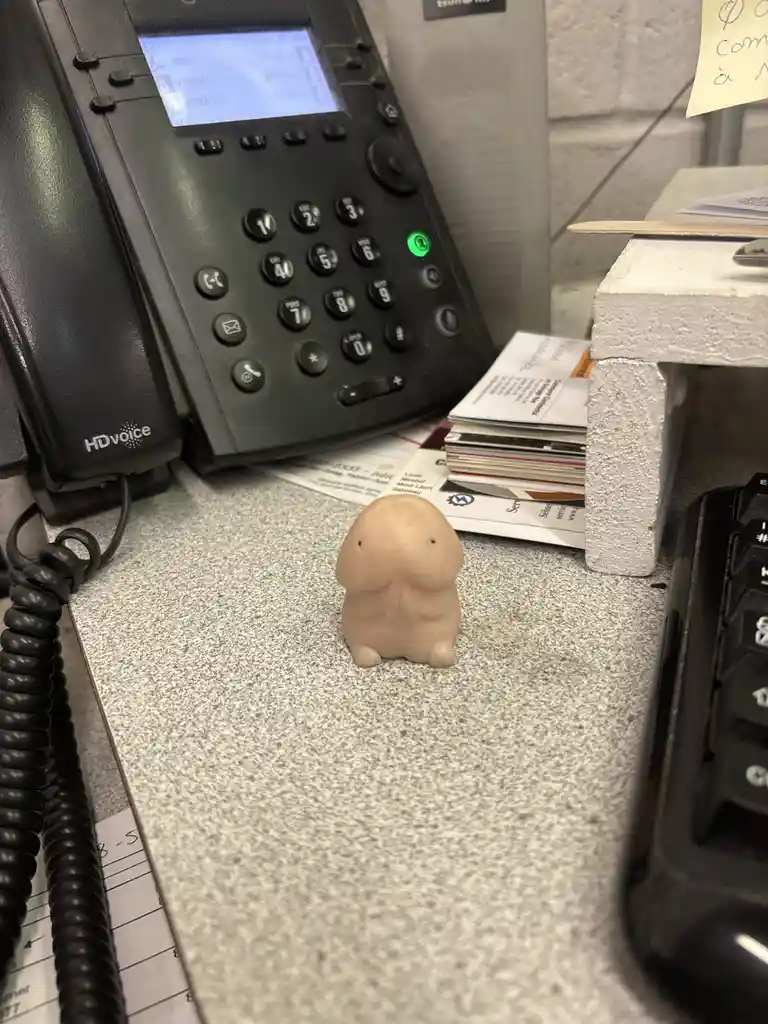 A small, beige, rabbit-like figurine sits on a speckled office desk, surrounded by a black HDvoice office phone, business…