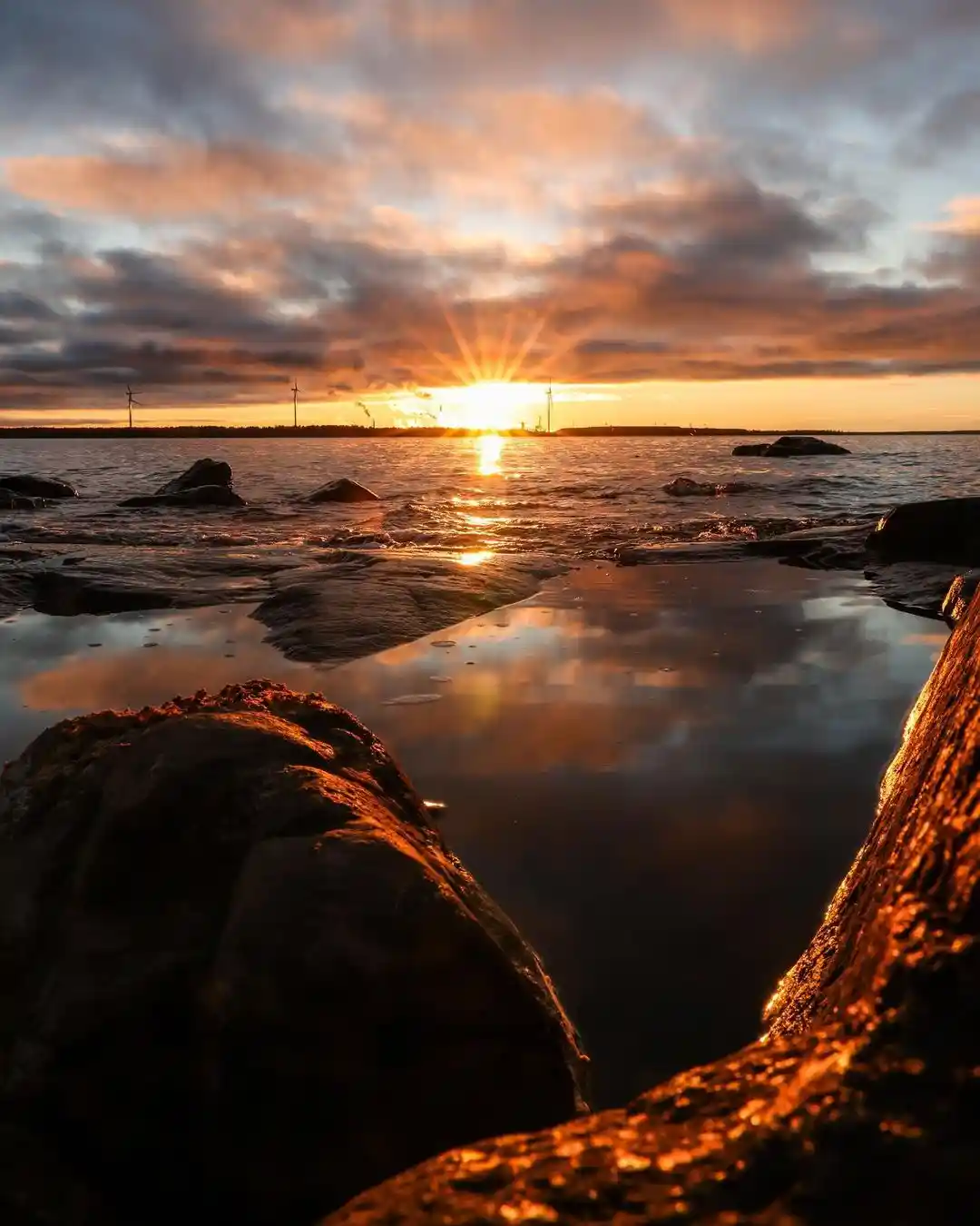 A serene sunset over a rocky shoreline with the sun low on the horizon, casting golden light across the water and clouds…