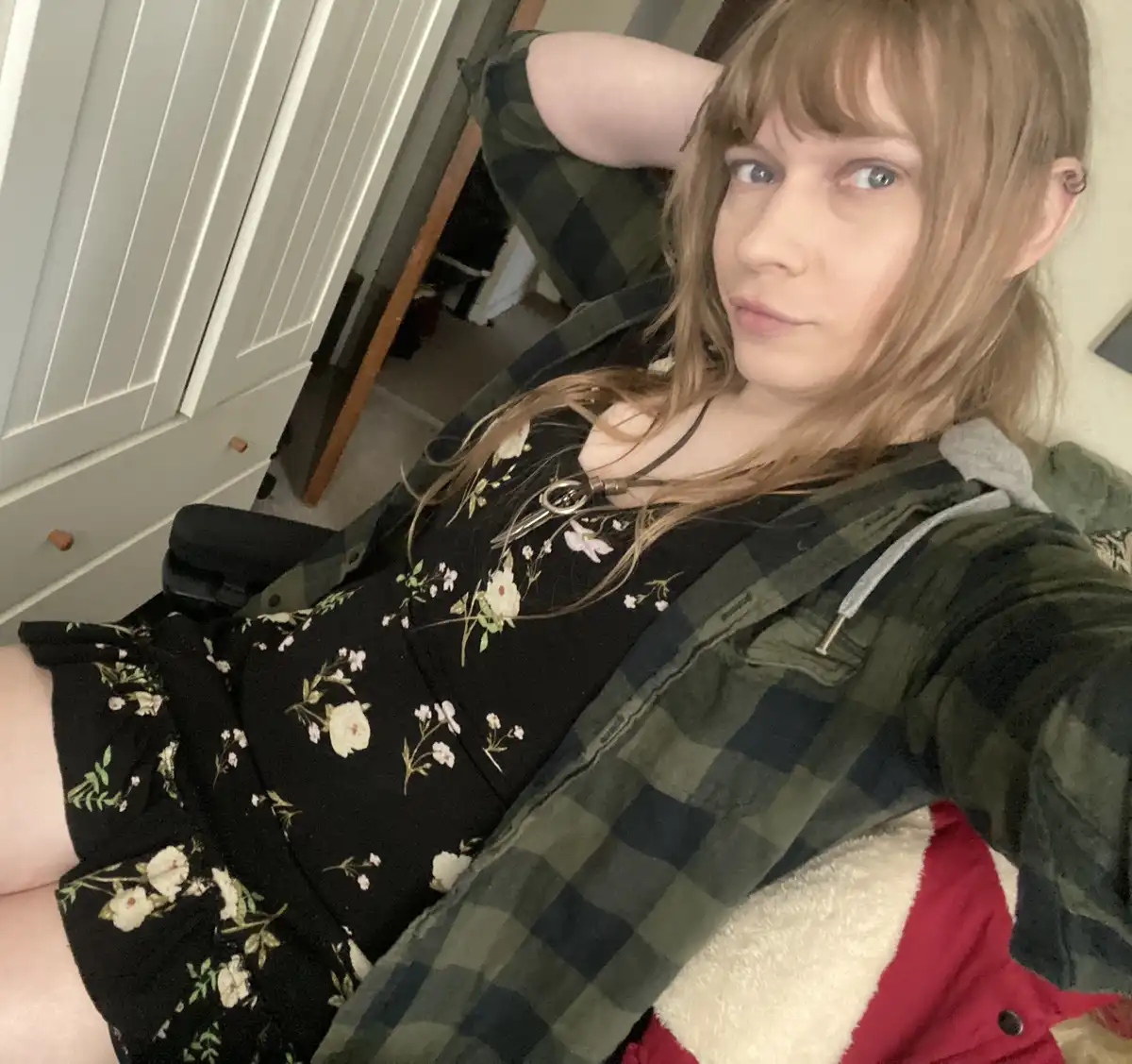 A person with light brown hair and blue eyes, wearing a black floral dress under an open green plaid shirt, lounges in a…