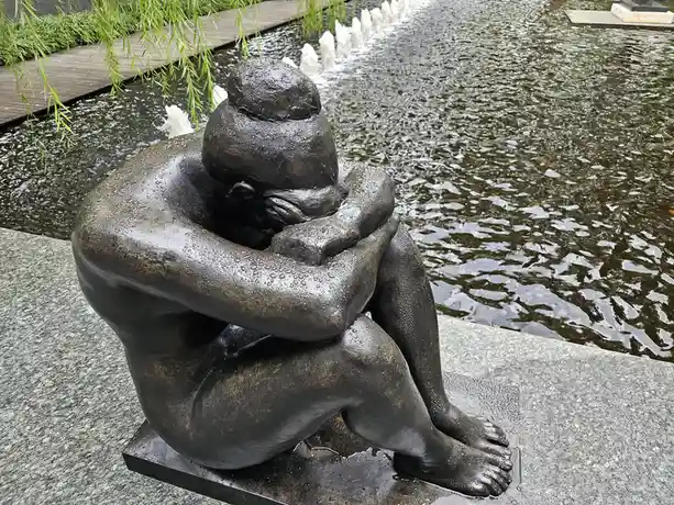 A bronze statue of a person sitting with their head buried in their arms, appearing to cry, positioned beside a water…
