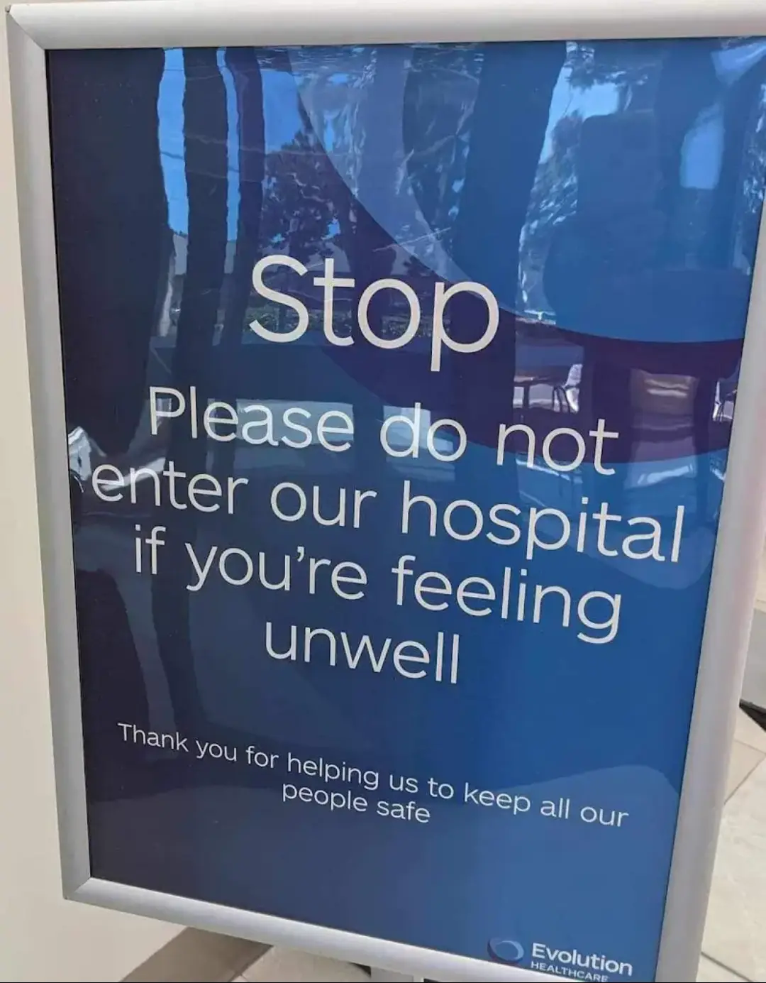 A blue hospital sign in a silver frame reads 'Stop Please do not enter our hospital if you're feeling unwell Thank you for…