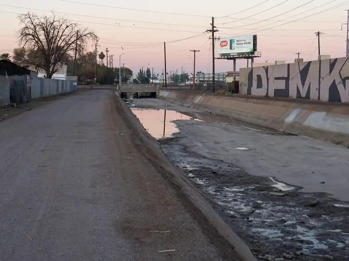A desolate urban drainage canal with muddy water and debris runs alongside a paved road, under a pink-hued twilight sky. A…