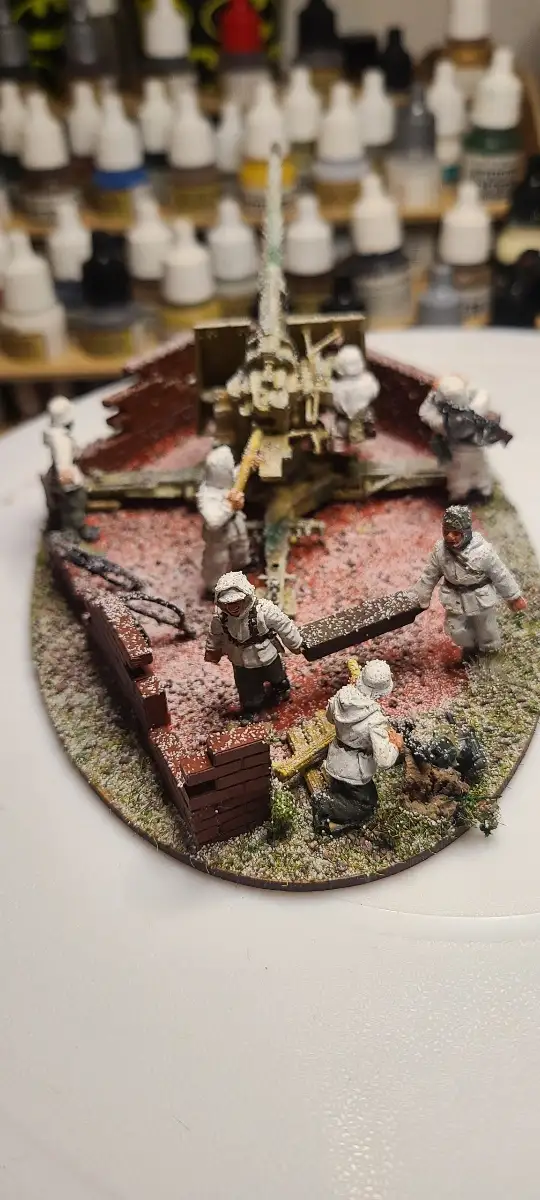 A close-up photo of a detailed miniature diorama featuring several painted soldier figures around a large artillery piece on…