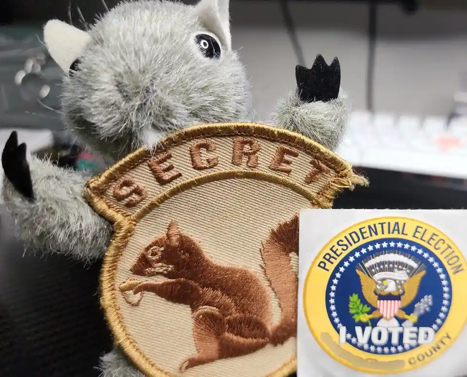 A plush gray squirrel holds a 'SECRET' patch featuring an embroidered squirrel holding a nut, next to a 'PRESIDENTIAL…