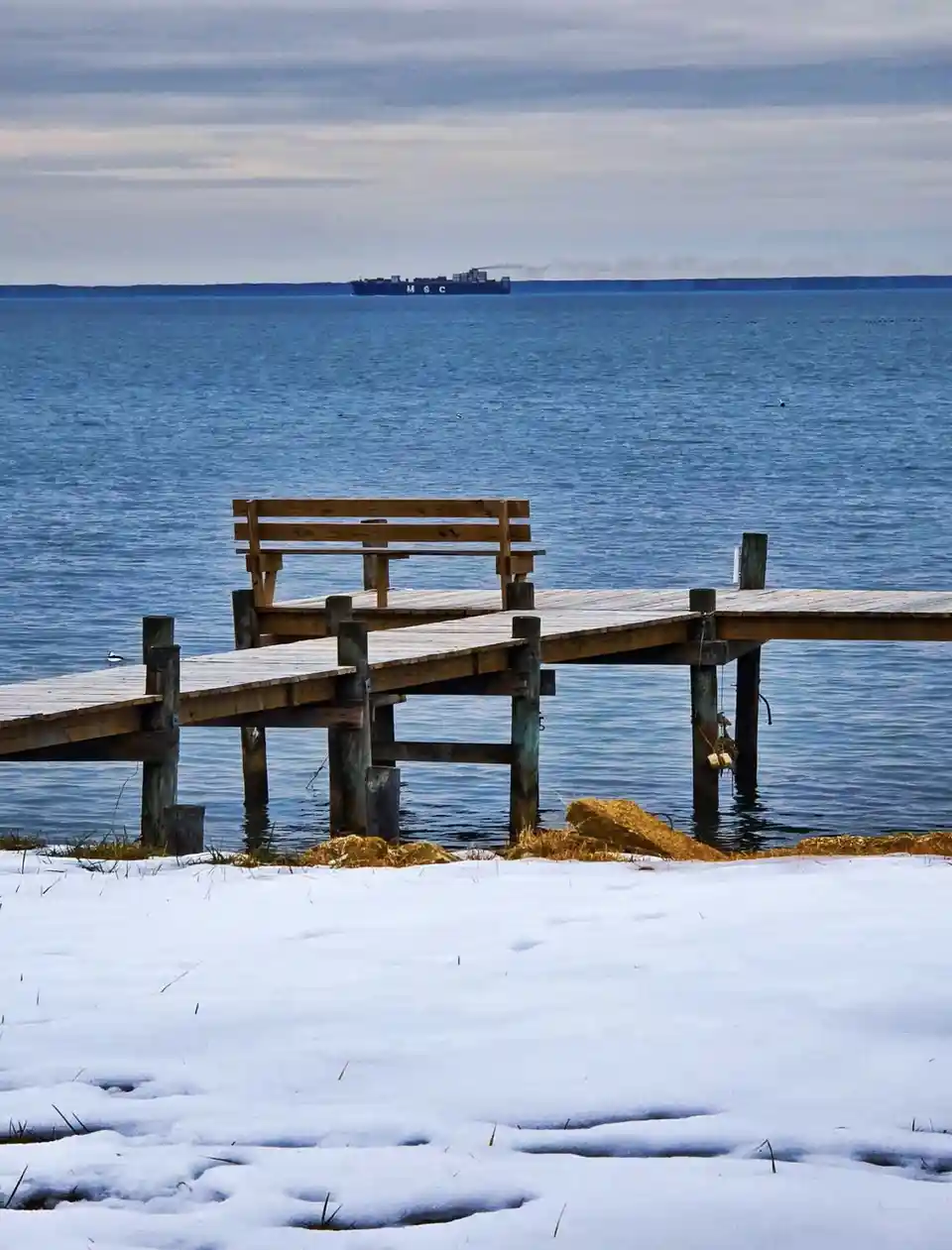 A snowy landscape features a wooden pier with a bench extending over calm blue water, with a large cargo ship labeled 'MSC'…