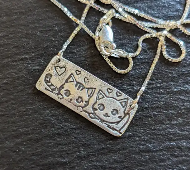 A silver-colored rectangular pendant necklace with two embossed cartoon cats and small hearts, attached to a box chain…