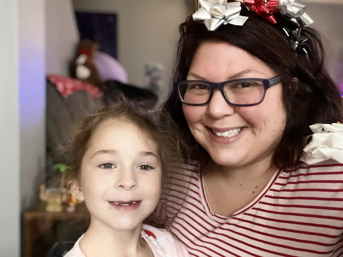 A woman and a young girl smile for the camera; the woman wears glasses and a headband with red, silver, and black bows…