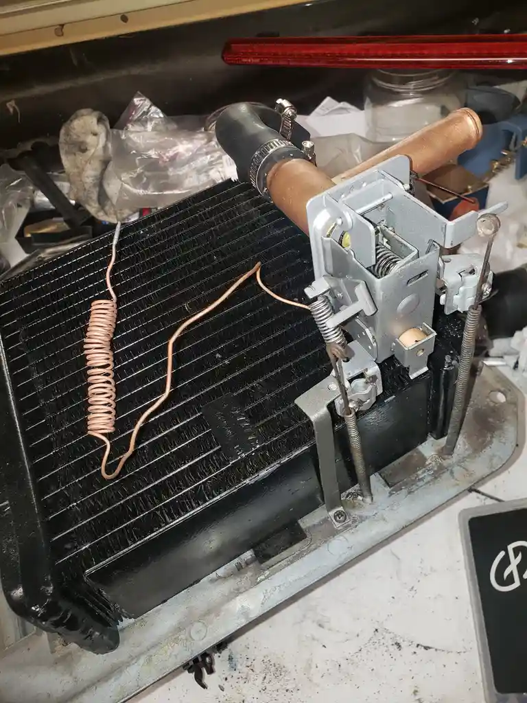 A close-up photo shows a car's heater core assembly with copper tubing and metal components, sitting on a workbench. A…