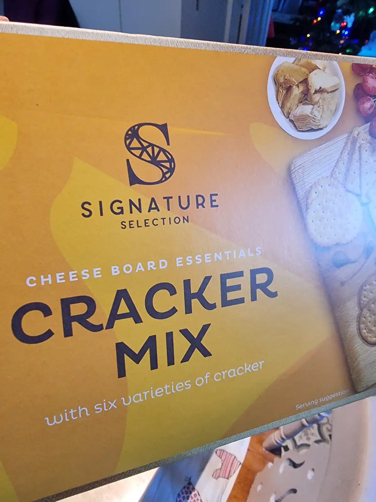 A close-up photo of a yellow box labeled 'Signature Selection Cheese Board Essentials Cracker Mix with six varieties of…